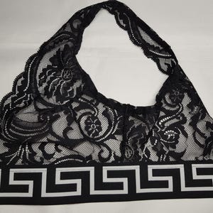 May include: Black lace bralette with a halter neck design. The bralette features intricate floral patterns and a decorative band with a Greek key design and a Medusa head emblem.