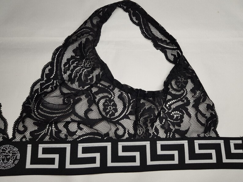May include: Black lace bralette with a halter neck design. The bralette features intricate floral patterns and a decorative band with a Greek key design and a Medusa head emblem.
