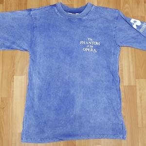 Puede incluir: Una camiseta azul desteñida con el texto "The Phantom of the Opera" impreso en la parte delantera. La camiseta tiene un aspecto descolorido y vintage.