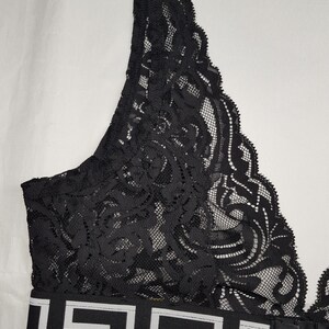 May include: Black lace bra with a decorative pattern. The bra features a wide band with a white and black geometric design. The bra has adjustable straps and a hook-and-eye closure.