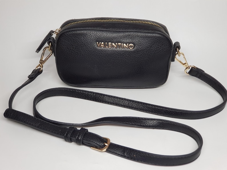 Pu&ograve; includere: Borsa a tracolla nera con cerniera dorata e tracolla lunga e regolabile. La borsa presenta il nome del marchio "VALENTINO" in lettere dorate sul davanti. La borsa &egrave; realizzata in materiale strutturato.