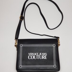 Puede incluir: Bolso bandolera negro con solapa rectangular. El texto "VERSACE JEANS COUTURE" en blanco est&aacute; enmarcado por un dise&ntilde;o de cuerda blanca. Tiene una correa larga y ajustable negra y detalles dorados.