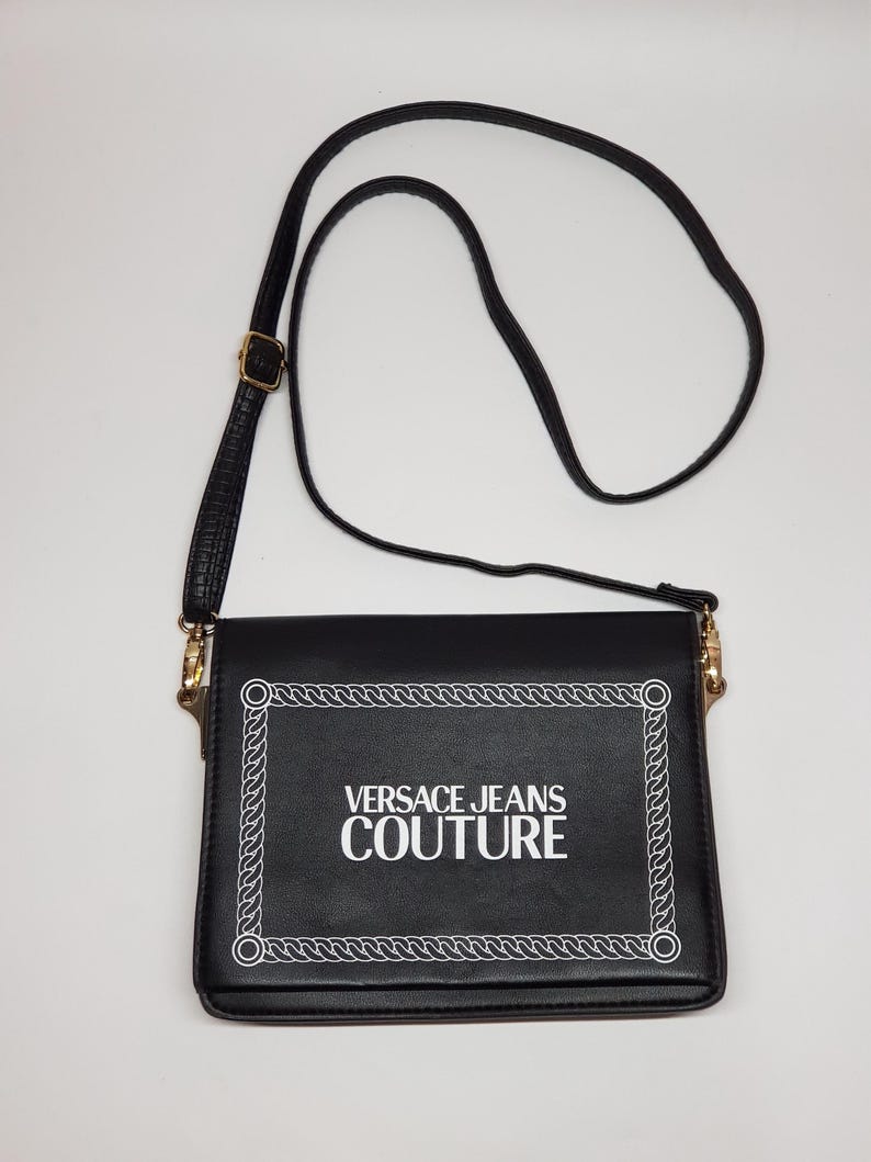 Puede incluir: Bolso bandolera negro con solapa rectangular. El texto "VERSACE JEANS COUTURE" en blanco est&aacute; enmarcado por un dise&ntilde;o de cuerda blanca. Tiene una correa larga y ajustable negra y detalles dorados.