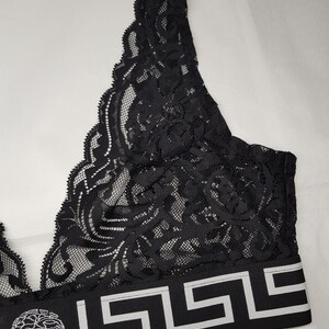 May include: Black lace bralette with a decorative floral pattern. The bralette features a wide band with a geometric design and a circular emblem. The band is black with white accents. The bralette is made of a delicate, sheer fabric.