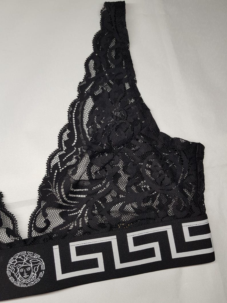 May include: Black lace bralette with a decorative floral pattern. The bralette features a wide band with a geometric design and a circular emblem. The band is black with white accents. The bralette is made of a delicate, sheer fabric.