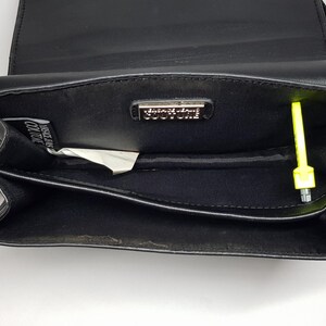 Puede incluir: Bolso bandolera de cuero negro con cierre de solapa. El interior presenta un peque&ntilde;o bolsillo y un logotipo met&aacute;lico. Un objeto amarillo ne&oacute;n est&aacute; dentro del bolso. El bolso tiene una correa de cadena dorada.