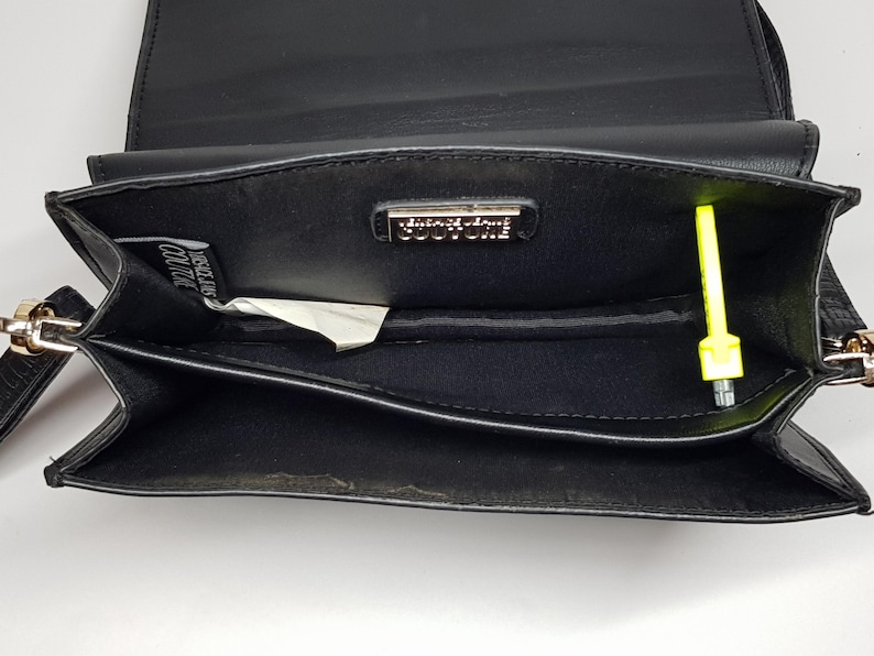 Puede incluir: Bolso bandolera de cuero negro con cierre de solapa. El interior presenta un peque&ntilde;o bolsillo y un logotipo met&aacute;lico. Un objeto amarillo ne&oacute;n est&aacute; dentro del bolso. El bolso tiene una correa de cadena dorada.