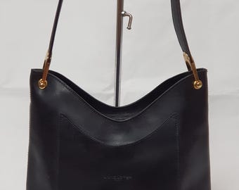 Könnte beinhalten: Eine schwarze Ledertasche mit rechteckiger Form und geschwungenem Oberteil. Die Tasche hat einen einzelnen Henkel und goldfarbene Beschläge. Der Markenname "LANCASTER" ist auf der Vorderseite sichtbar.