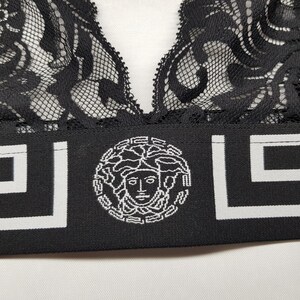 May include: Black lace bralette with a wide black band featuring a white Greek key design and a Medusa head emblem. The bralette has a delicate, floral lace pattern.