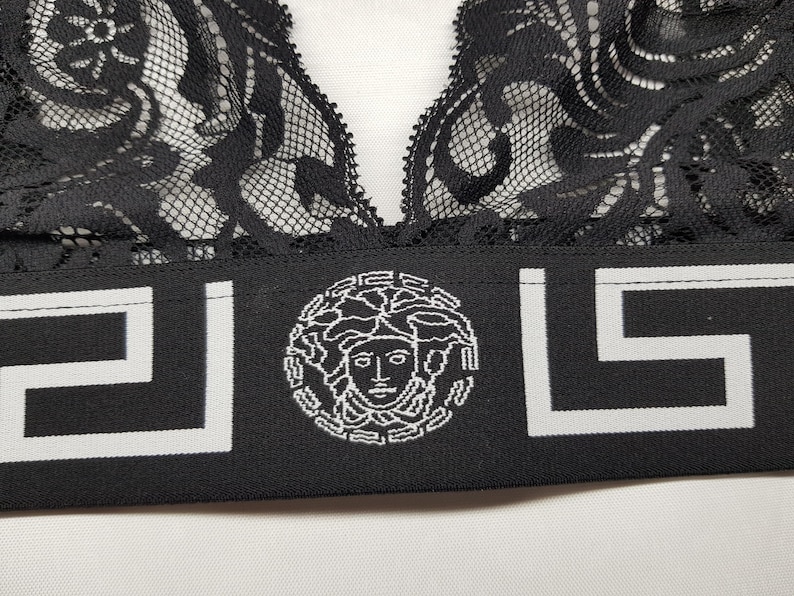 May include: Black lace bralette with a wide black band featuring a white Greek key design and a Medusa head emblem. The bralette has a delicate, floral lace pattern.