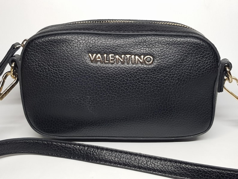 Pu&ograve; includere: Borsa a tracolla nera con finitura strutturata. La borsa presenta una cerniera e hardware dorati e il nome del marchio "VALENTINO" &egrave; visualizzato in lettere argentate sul davanti. Include una tracolla staccabile.