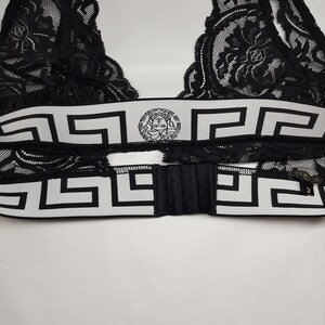May include: Black lace bralette with a white and black geometric patterned band. The band features a circular emblem and a hook-and-eye closure. The bralette is made of delicate lace.