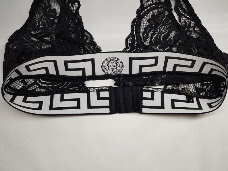 May include: Black lace bralette with a white and black geometric patterned band. The band features a circular emblem and a hook-and-eye closure. The bralette is made of delicate lace.