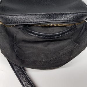 Pu&ograve; includere: Borsa a tracolla nera con esterno strutturato e cerniera dorata. La borsa ha un piccolo manico e una lunga tracolla. L'interno &egrave; in tessuto nero. La borsa &egrave; su una superficie bianca.