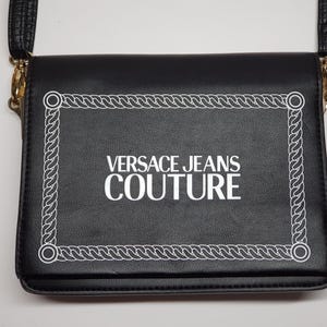 Puede incluir: Bolso bandolera negro con un borde blanco estilo cuerda y el texto "VERSACE JEANS COUTURE" en blanco. El bolso tiene una correa negra y herrajes dorados.