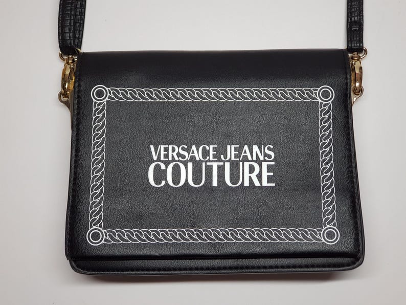 Puede incluir: Bolso bandolera negro con un borde blanco estilo cuerda y el texto "VERSACE JEANS COUTURE" en blanco. El bolso tiene una correa negra y herrajes dorados.