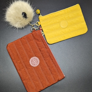 May include: Two small zippered pouches with a quilted design. One pouch is brown with a brown logo and the other is yellow with a brown logo. A fluffy white pom-pom keychain is attached to the brown pouch.