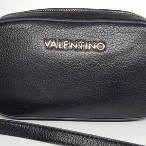 Pu&ograve; includere: Borsa a tracolla nera con aspetto in pelle strutturata. La borsa presenta una cerniera color argento e il nome del marchio "VALENTINO" in lettere argentate sul davanti. Un cinturino nero &egrave; visibile nella parte inferiore.