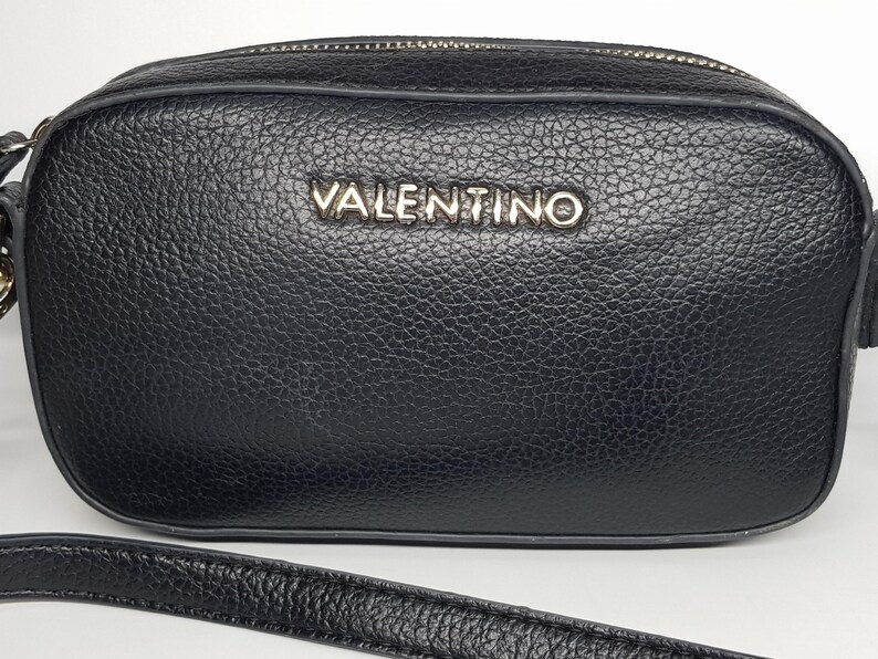 Pu&ograve; includere: Borsa a tracolla nera con aspetto in pelle strutturata. La borsa presenta una cerniera color argento e il nome del marchio "VALENTINO" in lettere argentate sul davanti. Un cinturino nero &egrave; visibile nella parte inferiore.