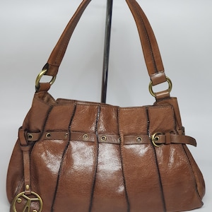 May include: A brown leather handbag with a curved top handle and a shoulder strap. The bag features a series of vertical seams and decorative metal hardware. A gold-toned charm hangs from the front.