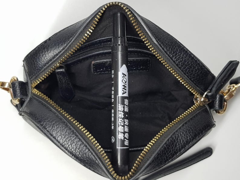 Pu&ograve; includere: Borsa a tracolla in pelle nera con cerniera e hardware dorati. All'interno della borsa c'&egrave; un pennarello nero con testo bianco "AOWA". La borsa ha un piccolo manico e una lunga tracolla.