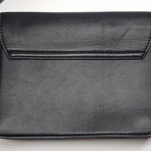 Puede incluir: Bolso bandolera negro con cierre de solapa. El bolso tiene una superficie texturizada y una correa de cadena dorada. Las costuras son visibles a lo largo de los bordes del bolso. El bolso es de forma rectangular.