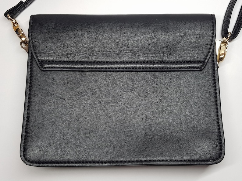 Puede incluir: Bolso bandolera negro con cierre de solapa. El bolso tiene una superficie texturizada y una correa de cadena dorada. Las costuras son visibles a lo largo de los bordes del bolso. El bolso es de forma rectangular.
