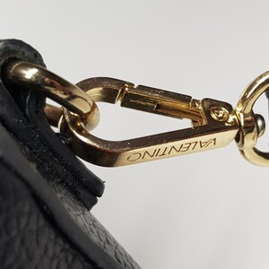Pu&ograve; includere: Primo piano di una chiusura in metallo color oro con il nome del marchio "VALENTINO" inciso. La chiusura &egrave; attaccata a un cinturino in pelle nera, che mostra un accessorio di lusso.