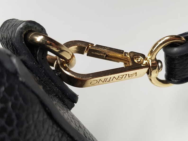 Pu&ograve; includere: Primo piano di una chiusura in metallo color oro con il nome del marchio "VALENTINO" inciso. La chiusura &egrave; attaccata a un cinturino in pelle nera, che mostra un accessorio di lusso.