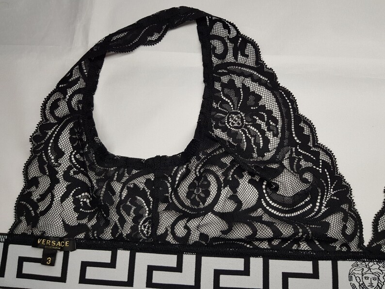 May include: Black lace halter top with intricate floral patterns. The top features a white and black geometric patterned band with the Versace logo and size 3. The top is made of a delicate, see-through lace material.