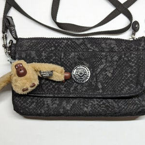 Peut inclure: Sac à bandoulière noir avec un motif peau de serpent et un bouton argenté. Un porte-clés singe brun est attaché au sac.