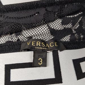 May include: Close-up of black lace underwear with the "VERSACE UNDERWEAR" label in gold lettering. The size tag reads "3". The background features a black and white geometric pattern.