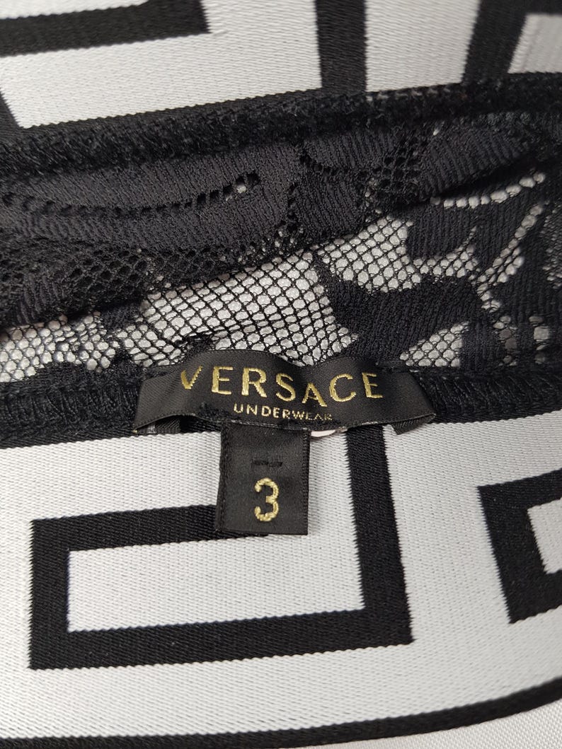May include: Close-up of black lace underwear with the "VERSACE UNDERWEAR" label in gold lettering. The size tag reads "3". The background features a black and white geometric pattern.