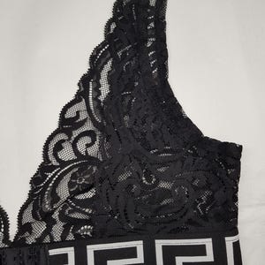 May include: Black lace bralette with a decorative pattern. The bralette features a wide band with a white and black geometric design. The straps and back closure are black.