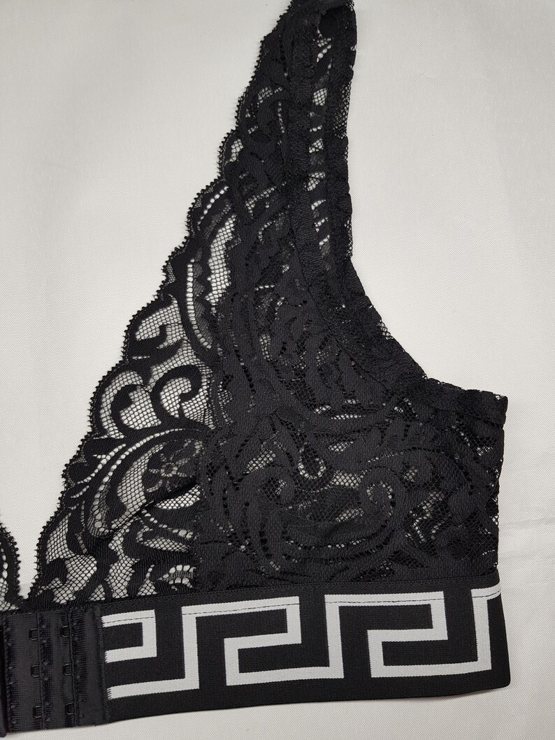 May include: Black lace bralette with a decorative pattern. The bralette features a wide band with a white and black geometric design. The straps and back closure are black.