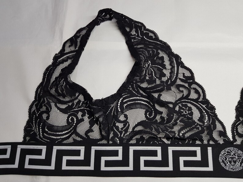 May include: Black lace halter top with a decorative pattern. The top features a black and white geometric band at the base with a Medusa head emblem. The top is designed with a halter neck and a delicate, intricate lace design.