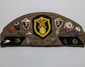 May include: A green military cap with a variety of pins and patches, including a red star, a tank, and a number "2".