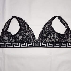 May include: Black lace bralette with a halter neck design. The bralette features intricate floral patterns and a band with a Greek key design and a circular emblem. The garment is displayed on a white background.