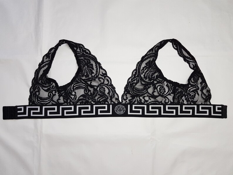 May include: Black lace bralette with a halter neck design. The bralette features intricate floral patterns and a band with a Greek key design and a circular emblem. The garment is displayed on a white background.
