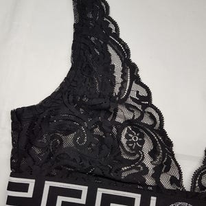 May include: Black lace bralette with a white geometric pattern on the band. The bralette features a delicate floral design and a Medusa head logo on the band. The bralette is made of a soft, sheer material.