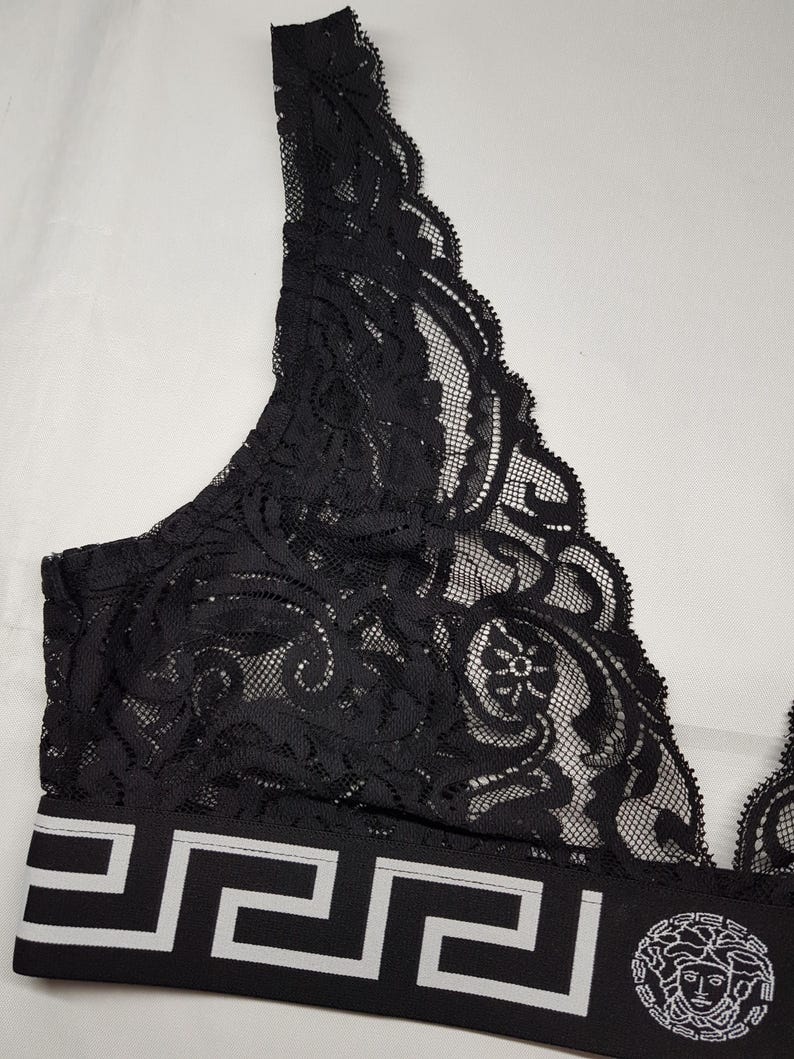 May include: Black lace bralette with a white geometric pattern on the band. The bralette features a delicate floral design and a Medusa head logo on the band. The bralette is made of a soft, sheer material.