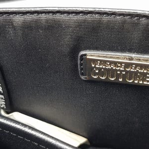 Puede incluir: Primer plano de una tela negra con dos etiquetas. Una etiqueta dice "VERSACE JEANS COUTURE" en texto blanco. La otra etiqueta es un rect&aacute;ngulo dorado con las palabras "VERSACE JEANS COUTURE" en texto negro.