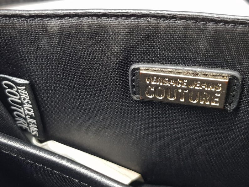 Puede incluir: Primer plano de una tela negra con dos etiquetas. Una etiqueta dice "VERSACE JEANS COUTURE" en texto blanco. La otra etiqueta es un rect&aacute;ngulo dorado con las palabras "VERSACE JEANS COUTURE" en texto negro.