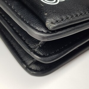 Puede incluir: Primer plano de una cartera de cuero negro con un dise&ntilde;o de cuerda blanca en la solapa. La cartera tiene un dise&ntilde;o en capas con costuras visibles y esquinas redondeadas. El fondo es blanco.