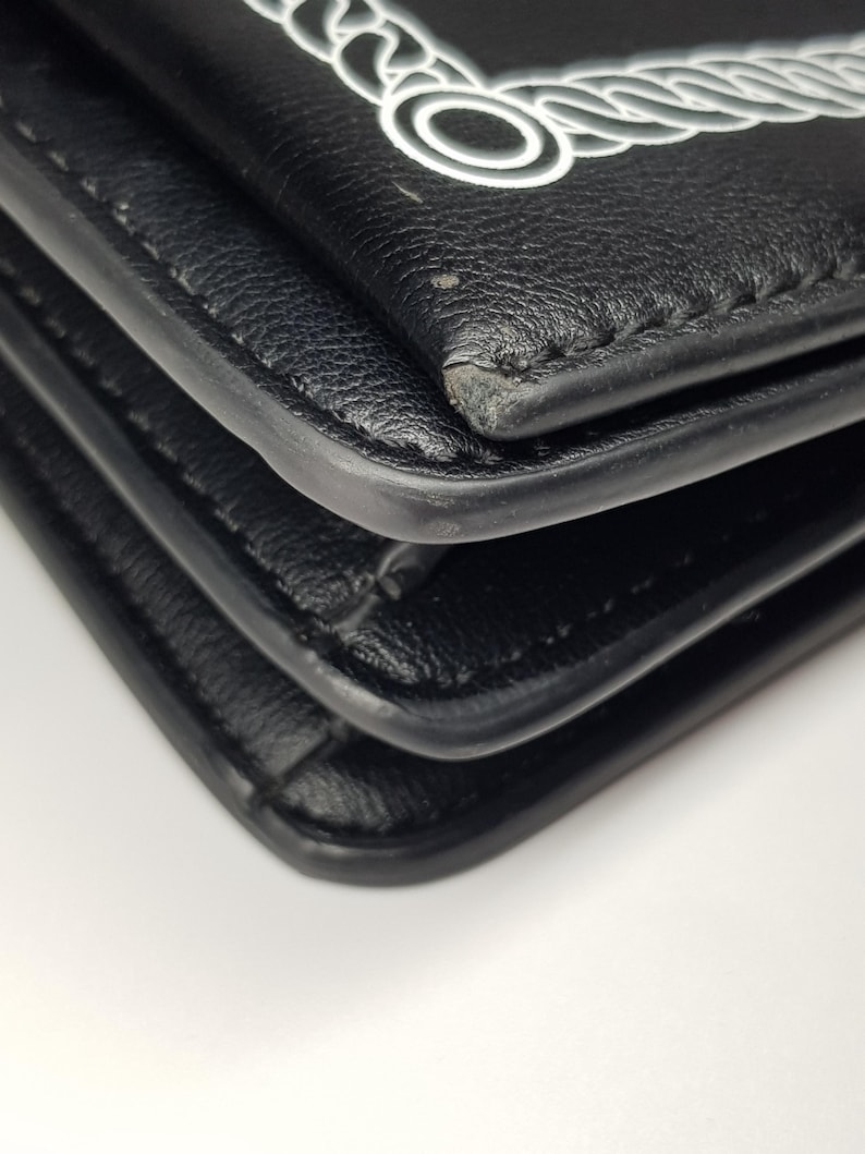 Puede incluir: Primer plano de una cartera de cuero negro con un dise&ntilde;o de cuerda blanca en la solapa. La cartera tiene un dise&ntilde;o en capas con costuras visibles y esquinas redondeadas. El fondo es blanco.