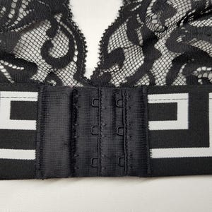 May include: Black lace bralette with a decorative pattern. The bralette features a black band with a white geometric design and a hook-and-eye closure. The lace is intricate and detailed, creating a delicate and feminine look.