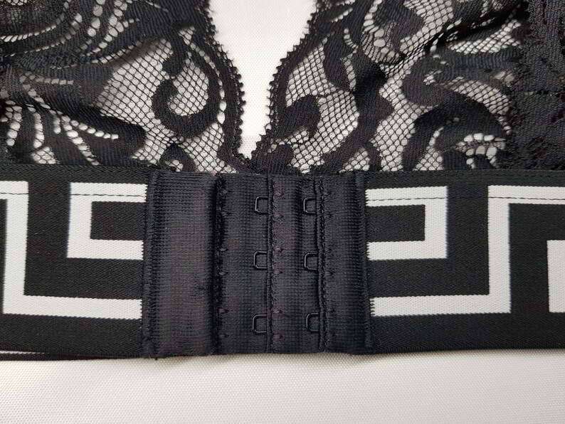 May include: Black lace bralette with a decorative pattern. The bralette features a black band with a white geometric design and a hook-and-eye closure. The lace is intricate and detailed, creating a delicate and feminine look.