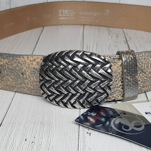 May include: A distressed leather belt with a silver-colored, woven oval buckle. The belt is a light tan color with a textured appearance. A tag with text is visible near the buckle.