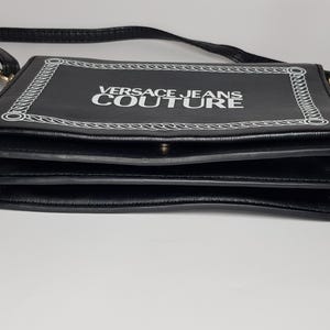 Puede incluir: Bolso bandolera negro de forma rectangular. El bolso presenta el texto "VERSACE JEANS COUTURE" en blanco, enmarcado por un dise&ntilde;o de cadena blanca. El bolso tiene una correa negra y un cierre dorado.
