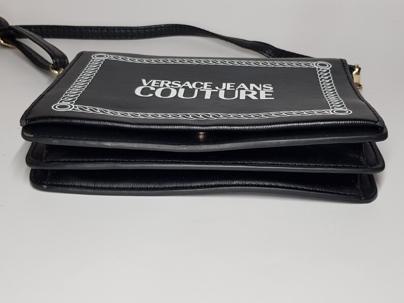Puede incluir: Bolso bandolera negro de forma rectangular. El bolso presenta el texto "VERSACE JEANS COUTURE" en blanco, enmarcado por un dise&ntilde;o de cadena blanca. El bolso tiene una correa negra y un cierre dorado.